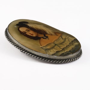 Puede incluir: Broche ovalado vintage con un retrato pintado a mano de una mujer con sombrero y pluma. El retrato est&aacute; engastado en un marco de metal plateado con un dise&ntilde;o de cuerda trenzada. La mujer lleva un vestido con escote con volantes.