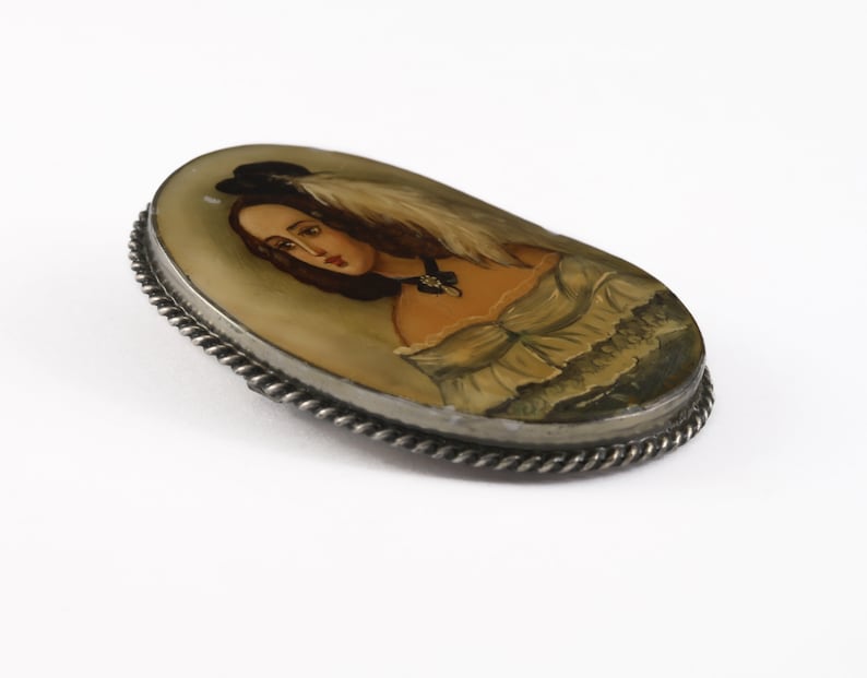 Puede incluir: Broche ovalado vintage con un retrato pintado a mano de una mujer con sombrero y pluma. El retrato est&aacute; engastado en un marco de metal plateado con un dise&ntilde;o de cuerda trenzada. La mujer lleva un vestido con escote con volantes.