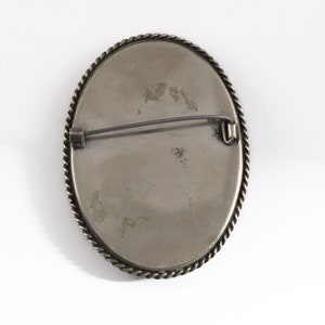 Puede incluir: Broche ovalado plateado con un borde trenzado en forma de cuerda. El broche tiene un cierre de alfiler en la parte posterior. El centro tiene una superficie gris claro con algo de decoloraci&oacute;n.