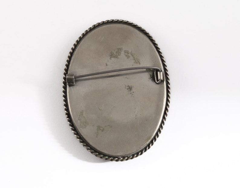 Puede incluir: Broche ovalado plateado con un borde trenzado en forma de cuerda. El broche tiene un cierre de alfiler en la parte posterior. El centro tiene una superficie gris claro con algo de decoloraci&oacute;n.