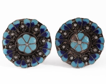 May include: A pair of round silver earrings with blue and turquoise enamel. The earrings feature a floral design with a central flower surrounded by smaller flowers.