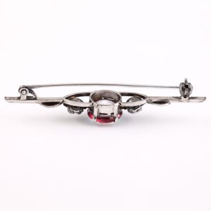 May include: A silver brooch with a red gemstone surrounded by smaller white gemstones. The brooch has a delicate floral design and a pin and clasp closure.