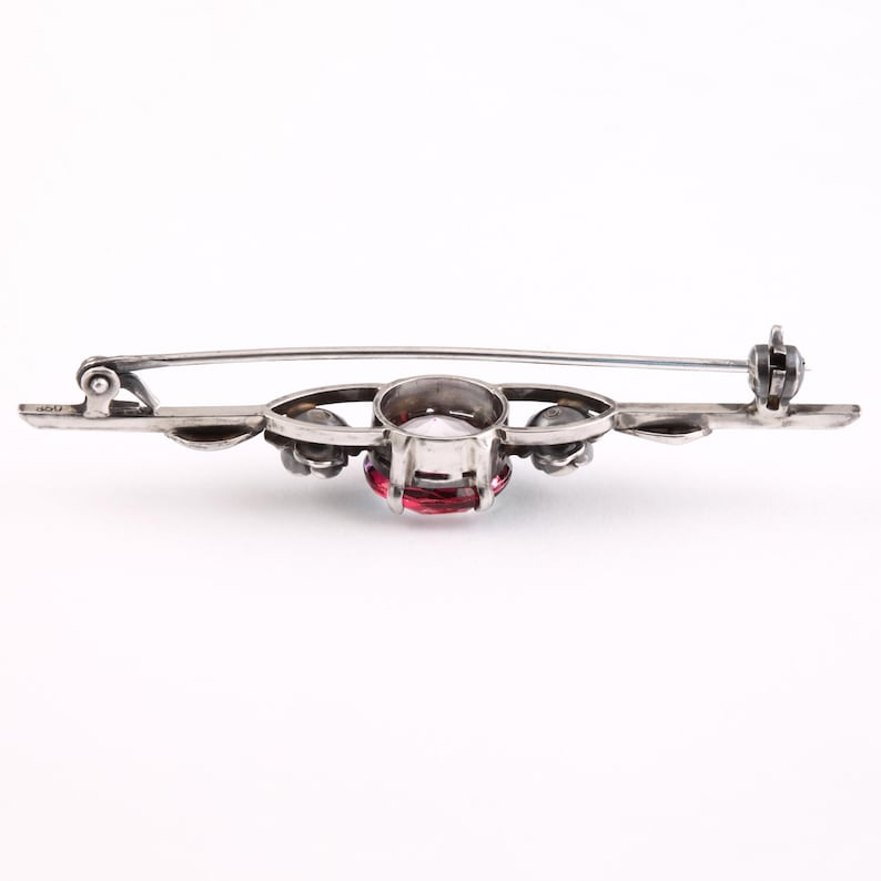 May include: A silver brooch with a red gemstone surrounded by smaller white gemstones. The brooch has a delicate floral design and a pin and clasp closure.