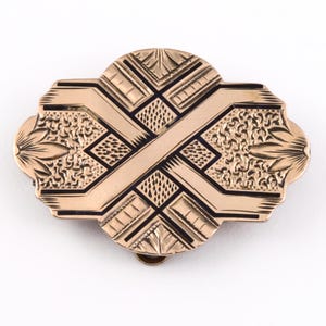 May include: A gold-toned brooch with a geometric design. The brooch has a black enamel inlay and a detailed engraved pattern.