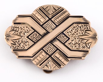 May include: A gold-toned brooch with a geometric design. The brooch has a black enamel inlay and a detailed engraved pattern.