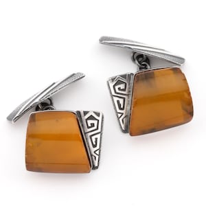 May include: A pair of cufflinks featuring square amber-colored stones with a silver-tone metal frame and backing. The silver metal has a textured finish and a decorative geometric design on the side of the amber stone.