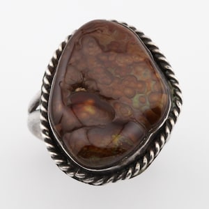 May include: A silver ring featuring a large, oval-shaped stone with a unique pattern of brown, tan, and green hues. The stone is set in a silver bezel with a twisted rope design around the edge. The ring has a simple, polished band.