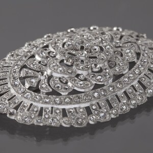 K&ouml;nnte beinhalten: Eine ovale Brosche aus Silber mit einem detaillierten Blumendesign und klaren Strasssteinen.