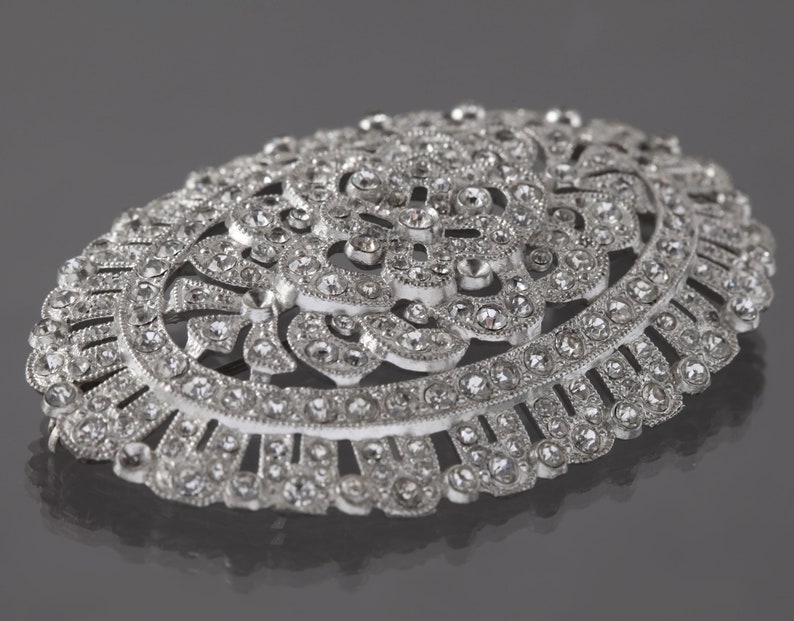 K&ouml;nnte beinhalten: Eine ovale Brosche aus Silber mit einem detaillierten Blumendesign und klaren Strasssteinen.