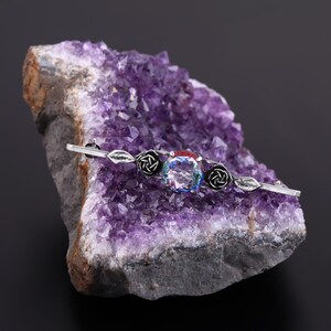 May include: A silver brooch with a large, clear gemstone surrounded by black enamel roses and leaves. The brooch is resting on a purple amethyst geode.