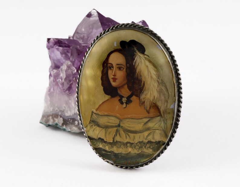 Puede incluir: Broche ovalado antiguo con un retrato de una mujer con vestido blanco y sombrero negro con pluma. El broche tiene un borde plateado en forma de cuerda. Un cristal de amatista p&uacute;rpura en el fondo.