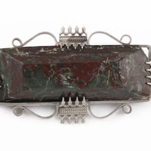 May include: A silver brooch with a rectangular green and red stone set in the center. The brooch has a decorative design with a series of small points around the stone.