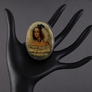Puede incluir: Broche ovalado vintage con un retrato pintado a mano de una mujer con vestido blanco y sombrero negro con pluma. El broche tiene un borde plateado retorcido. La mujer del retrato tiene cabello casta&ntilde;o y lleva un collar negro.