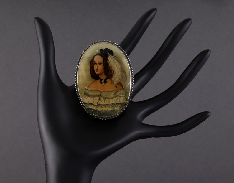 Puede incluir: Broche ovalado vintage con un retrato pintado a mano de una mujer con vestido blanco y sombrero negro con pluma. El broche tiene un borde plateado retorcido. La mujer del retrato tiene cabello casta&ntilde;o y lleva un collar negro.