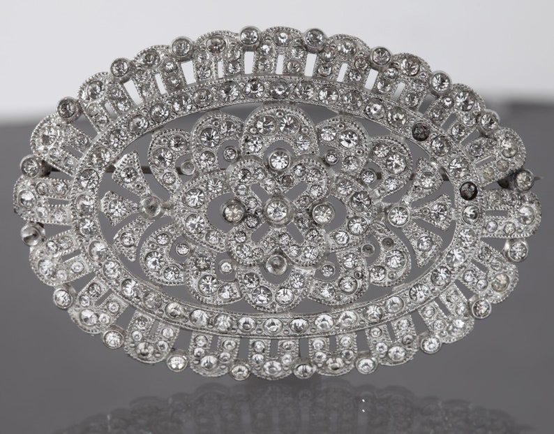 K&ouml;nnte beinhalten: Eine ovale Brosche aus Silber mit einem floralen Design, besetzt mit klaren Strasssteinen.