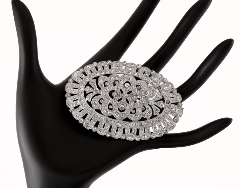 K&ouml;nnte beinhalten: Eine ovale Brosche aus Silber mit einem floralen Design, besetzt mit klaren Strasssteinen.
