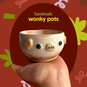 Op de afbeelding: Een kleine, witte keramische pot met een cartoon gezicht. De pot heeft een lichtbruine rand en gele, oranje en groene details. De pot wordt in een hand gehouden. De tekst "handmade wonky pots" is zichtbaar op de achtergrond.