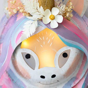 May include: A whimsical unicorn head with a golden horn, adorned with a white flower and dried floral accents. The unicorn's face is painted in soft pastel colors, with a glowing light emanating from within.