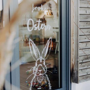 May include: A glass window featuring the words "frohe Ostern" in white script, alongside a white rabbit illustration. The window is framed in grey, and a wooden wall is visible to the right. The image has a festive, Easter theme.