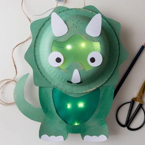 May include: A handmade green dinosaur lantern with white accents and glowing lights. The dinosaur is crafted from paper plates and cardstock, featuring a triceratops design. A pencil and scissors are visible in the background.