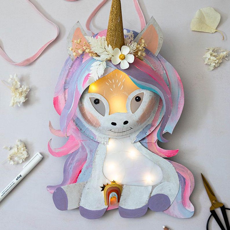 May include: A whimsical, handmade unicorn-shaped light fixture. The unicorn features a gold glitter horn, pastel-colored mane, and a rainbow accent. It has a string of small lights and is decorated with flowers. The light fixture is designed for wall hanging.