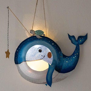 Puede incluir: Lámpara artesanal con forma de ballena, con una cara sonriente, cuerpo azul y vientre blanco. Una pequeña tortuga está encima. Una estrella dorada cuelga de una cuerda. Ideal para la habitación de un niño.