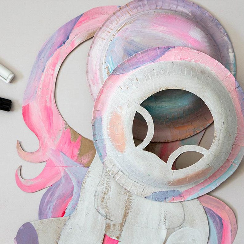 May include: A whimsical unicorn mask crafted from paper plates, painted with pastel pink, purple, and blue hues. The mask features cut-out eye holes and a white base, with a flowing mane and horn details. A creative craft project.