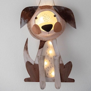 May include: A whimsical dog-shaped wall decoration with a warm, glowing interior. The dog is crafted from light-colored wood with brown accents on the ears, tail, and spots. The eyes, nose, and mouth are black. The interior has small lights.