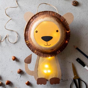 May include: A homemade lion lantern crafted from cardboard and paper. The lion features a brown mane, a yellow face, and a black nose. Small, warm white lights illuminate the lantern, producing a gentle glow. A fun craft project for children.
