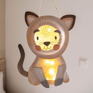 Può includere: Una lampada a sospensione a forma di gatto realizzata in legno con viso e corpo di colore chiaro. Il design presenta orecchie, coda e zampe dipinte in marrone scuro, con una calda luce interna che illumina il viso e il petto del gatto.