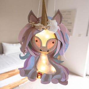 May include: A whimsical unicorn-shaped hanging lamp with a golden horn, adorned with pastel-colored paper mane and tail. The lamp features a warm, glowing light, creating a soft ambiance. The unicorn has a floral crown and is suspended by a thin, gold-colored string.