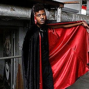 May include: A person wearing a black velvet cape with a red satin lining. The cape has a hood and is worn over a black shirt and pants.