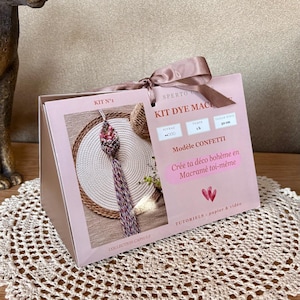 Puede incluir: Una caja rectangular de kit de manualidades de color rosa claro con un asa de cinta marrón. La caja incluye texto como KIT N°1, KIT DYE MACRAME y Modèle CONFETTI, junto con una imagen de un macramé. La caja indica un tamaño final de 50 cm.