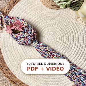 Peut inclure: Une suspension en macramé multicolore composée d'une petite pochette contenant des fleurs artificielles roses et d'un long pompon tressé. L'objet comporte une étiquette rectangulaire blanche avec le texte TUTORIEL NUMÉRIQUE PDF + VIDÉO en brun.