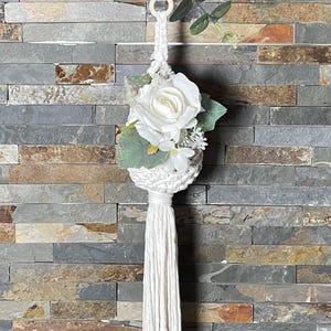 May include: A white macrame hanging with a white flower and green leaves. The hanging is attached to a wooden ring and has a long white tassel.
