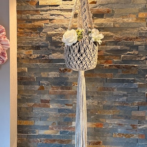Peut inclure: Un porte-plante en macramé de couleur crème avec un pot noir rempli de fleurs blanches. Le support est suspendu au plafond et possède une longue gland. Le fond est un mur de pierre, et une plante verte est visible en bas.