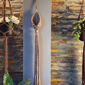 Peut inclure: Suspension de plante en macramé marron avec un motif de panier tissé et de longues franges tressées. Le support est suspendu contre un mur en pierre, mettant en valeur un style bohème. Pour l'intérieur ou l'extérieur.
