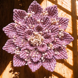 May include: A handcrafted macrame flower wall hanging in pink and purple hues. The petals surround a centre of dried, cream-coloured flowers. The piece is displayed on a wooden surface, with sunlight creating shadows across the piece.