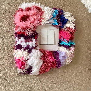 Può includere: Una placca quadrata per interruttore della luce decorata con un fitto ciuffo di lana testurizzata nei toni del rosa, magenta, bianco, viola e blu brillante. La parte centrale presenta un meccanismo a doppio interruttore bianco.