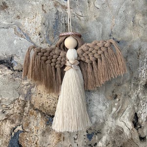 Op de afbeelding: Een handgemaakte macramé engel ornament. De engel heeft een houten kralen hoofd, een halo en vleugels gemaakt van geknoopt beige garen. Het lichaam is een crèmekleurige kwast, en de vleugels zijn donkerder beige. Een touwtje is bevestigd om op te hangen.