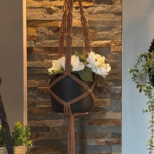 Peut inclure: Un porte-plante en macramé marron avec un pot noir rempli de fleurs blanches. Le support est suspendu à un anneau et est suspendu devant un mur de pierre. Le porte-plante est un objet décoratif pour les plantes d'intérieur.