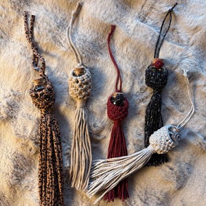 Peut inclure: Cinq désodorisants décoratifs pour voiture de différents modèles. L'un est en tissu imprimé léopard, un autre en macramé beige, un troisième bordeaux, un quatrième en paillettes noires et le dernier en macramé blanc avec fil argenté.