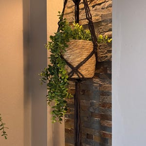 Peut inclure: Un porte-plante en macramé marron avec un panier tressé contenant de la verdure. Le support est suspendu à un luminaire de plafond. Le panier est beige et la verdure est verte. L'arrière-plan est un mur en pierre.