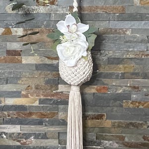 May include: A white macrame plant hanger with a white tassel and a bouquet of white flowers. The hanger is hanging from a wooden ring against a stone wall.