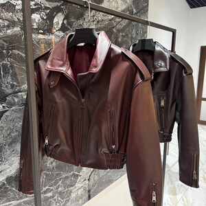 May include: A deep burgundy faux leather jacket featuring a wide collar, shoulder epaulets, a front zipper closure, and two vertical zippered pockets. The waist and sleeve cuffs are finished with a gathered elastic band detail.