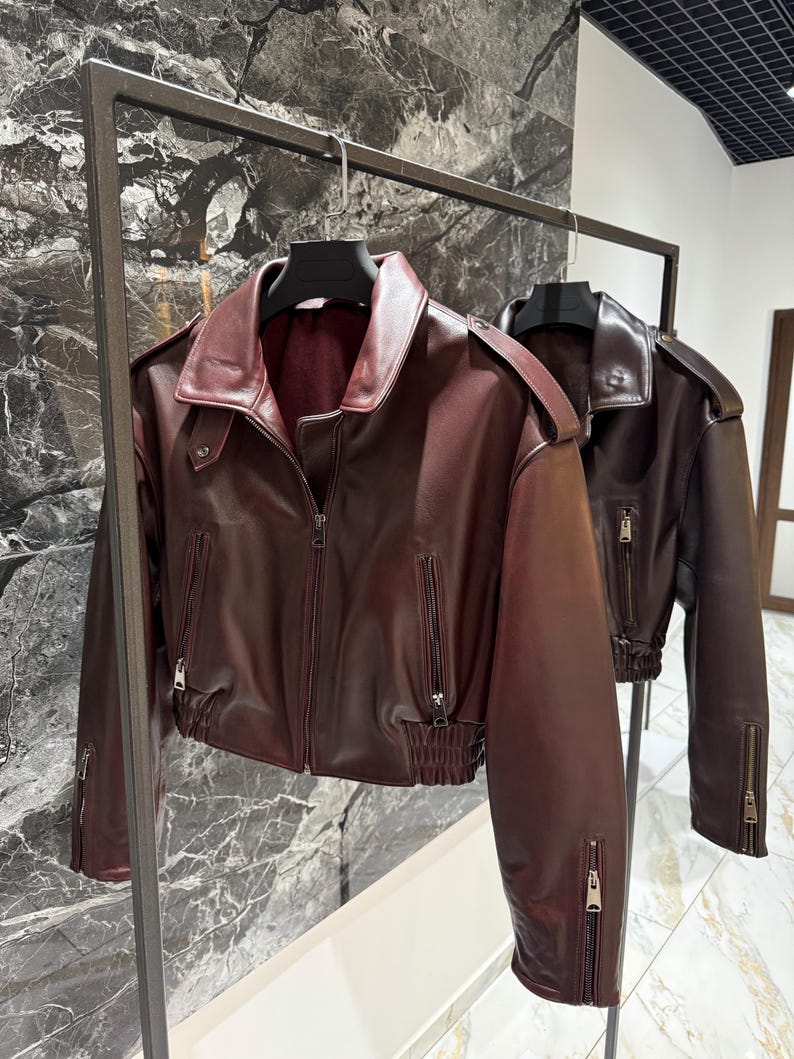 May include: A deep burgundy faux leather jacket featuring a wide collar, shoulder epaulets, a front zipper closure, and two vertical zippered pockets. The waist and sleeve cuffs are finished with a gathered elastic band detail.