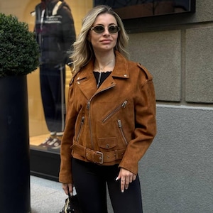 May include: A brown suede jacket with a belted waist, multiple zippered pockets, and shoulder epaulets. The jacket has a classic biker style and is worn with black pants. The jacket is a warm, earthy tone.