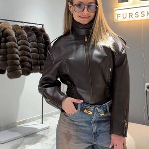 May include: A dark brown leather jacket with a high collar and zipper closure. The jacket has a fitted silhouette and is paired with blue jeans. A black belt with a gold buckle is visible.