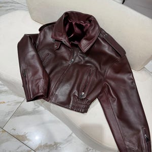 May include: A burgundy leather jacket with a classic collar, zipper closure, and zippered sleeves. The jacket has a fitted waist and shoulder epaulets. The interior lining is a darker shade of burgundy.