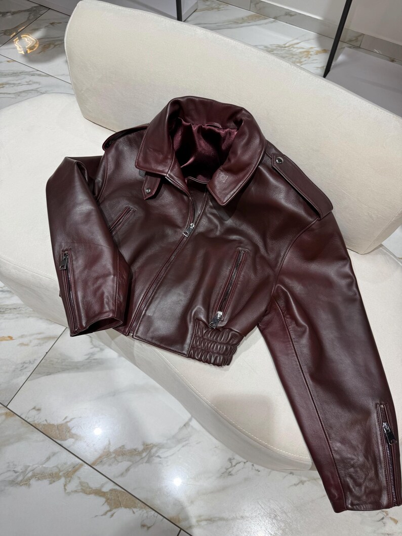May include: A burgundy leather jacket with a classic collar, zipper closure, and zippered sleeves. The jacket has a fitted waist and shoulder epaulets. The interior lining is a darker shade of burgundy.